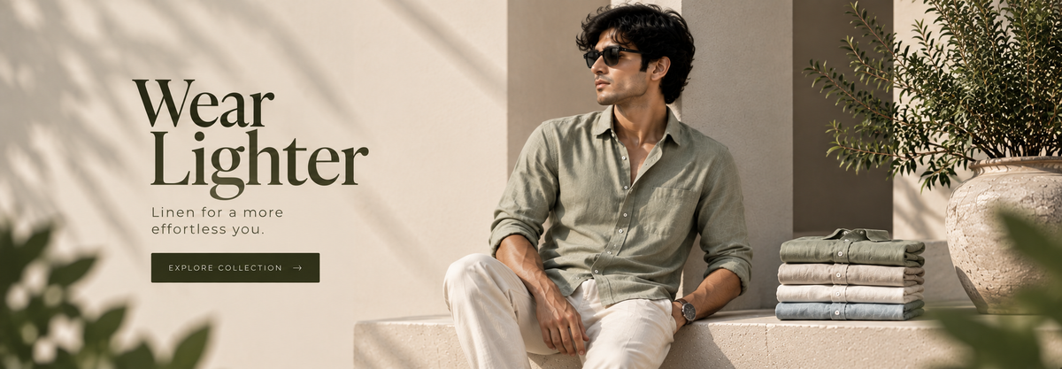 A man wearing a light sage green linen shirt and white trousers sits on a beige bench in a sunlit interior. The text 'Wear Lighter' appears on the left, with folded matching shirts stacked beside him and a potted plant on the right.